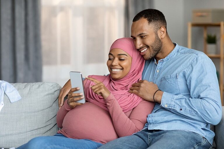 Pregnant woman and her partner looking at a smartphone together on a couch.