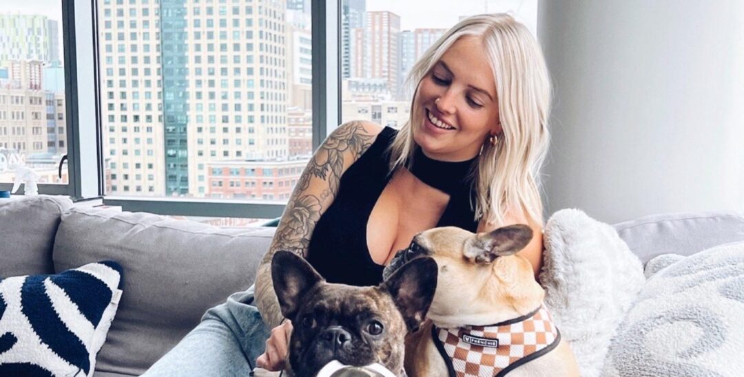 Blonde woman smiling while relaxing on a sofa with two French bulldogs.