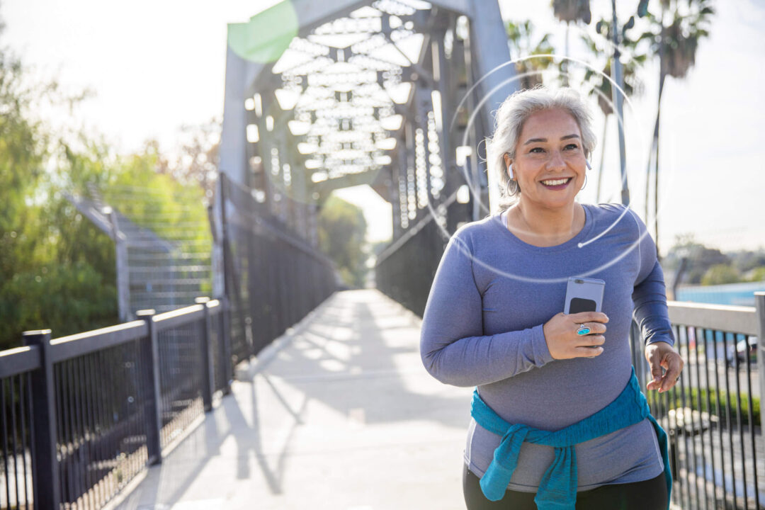 Smiling older woman jogging across a bridge while listening to music on her phone.