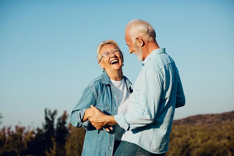 Senior couple holding hands and laughing together outdoors against a clear blue sky.