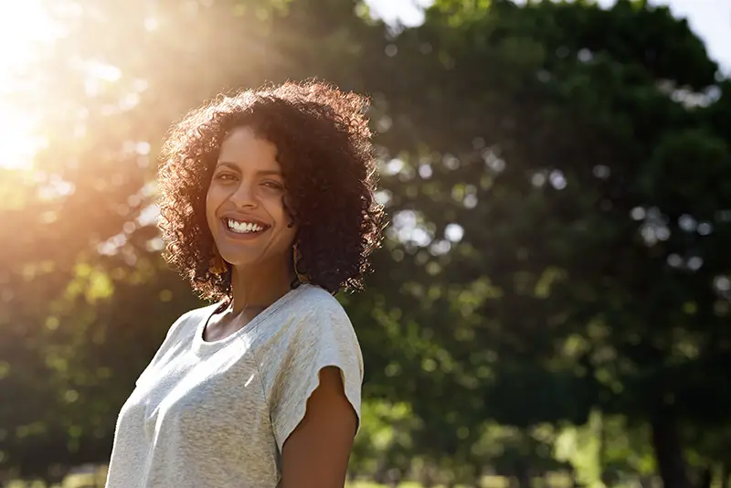 Smiling woman with curly hair standing outdoors in sunlight.