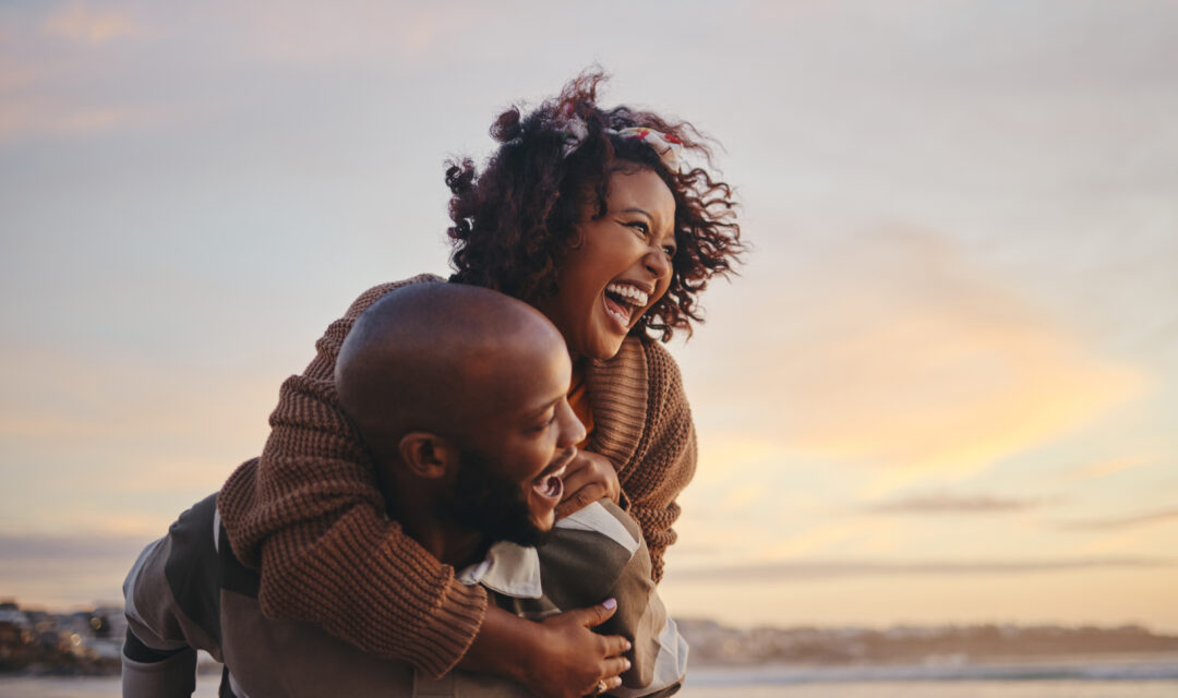 Man giving a woman a piggyback ride on a beach at sunset, both laughing.
