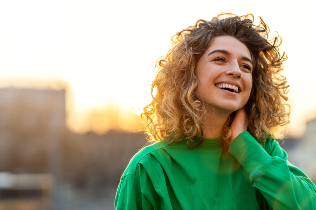 Smiling young woman with curly hair wearing a green top outdoors.