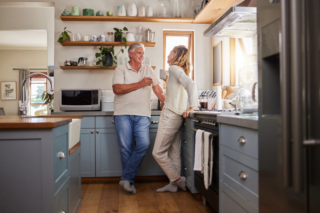 Mature couple talking and holding mugs while leaning on counters in a bright kitchen.